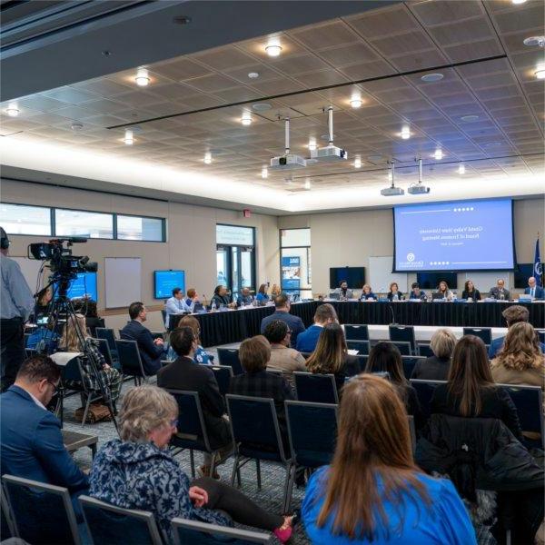 A group of people sit in chairs in a meeting room. Others are sitting at the head table. The screen says, "Grand Valley State University Board of Trustees meeting, February 21, 2025.